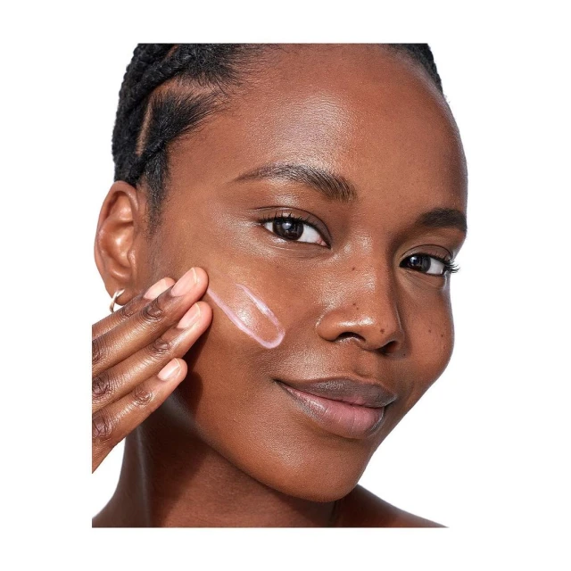 Close-up of a dark-skinned woman smiling as she applies a skincare cream to her cheek in a beauty advertisement shot.