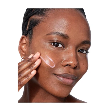 Close-up of a dark-skinned woman smiling as she applies a skincare cream to her cheek in a beauty advertisement shot.