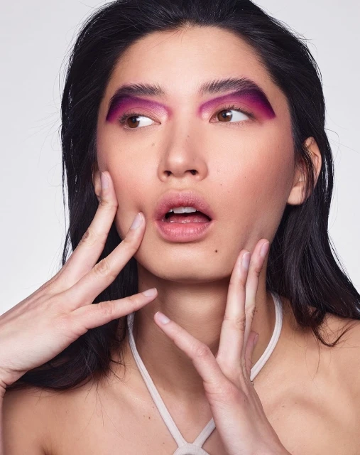 Close-up of an adult female model with makeup. She has dark hair, fair skin, and pink and purple makeup on her eyes and eyebrows.