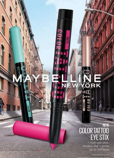 Maybelline Color Tattoo Eye Stix advertisement featuring three cosmetic products in different colors, buildings in the background.