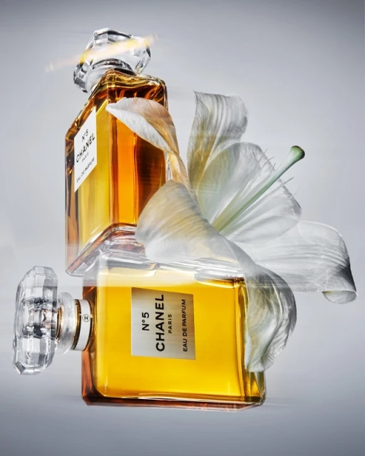 Two Chanel N°5 perfume bottles with golden liquid are stacked vertically and adorned with a white lily blossom, set against a gray backdrop.