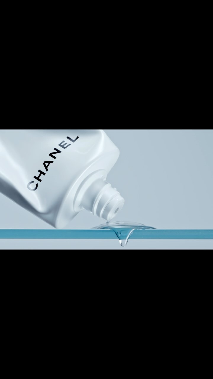 Close-up of a white Chanel skincare bottle dispensing a clear liquid onto a reflective surface against a soft blue backdrop.