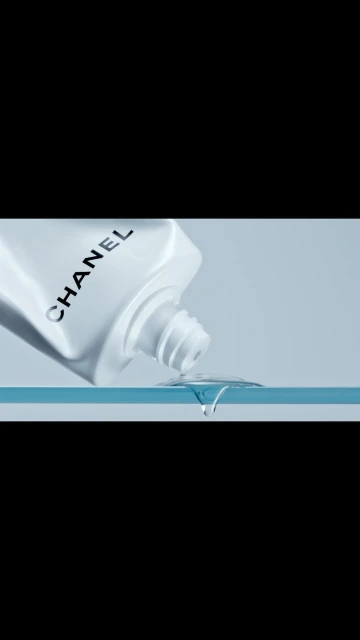 Close-up of a white Chanel skincare bottle dispensing a clear liquid onto a reflective surface against a soft blue backdrop.