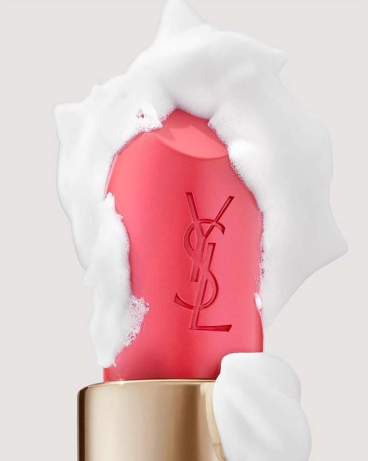 Close-up of a coral pink lipstick tube, partially covered in white foam, against a neutral background, highlighting beauty and cosmetic.