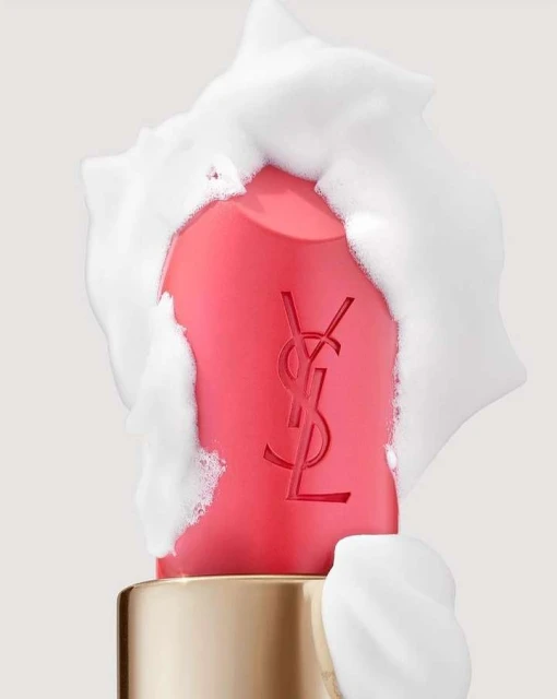 Close-up of a coral pink lipstick tube, partially covered in white foam, against a neutral background, highlighting beauty and cosmetic.
