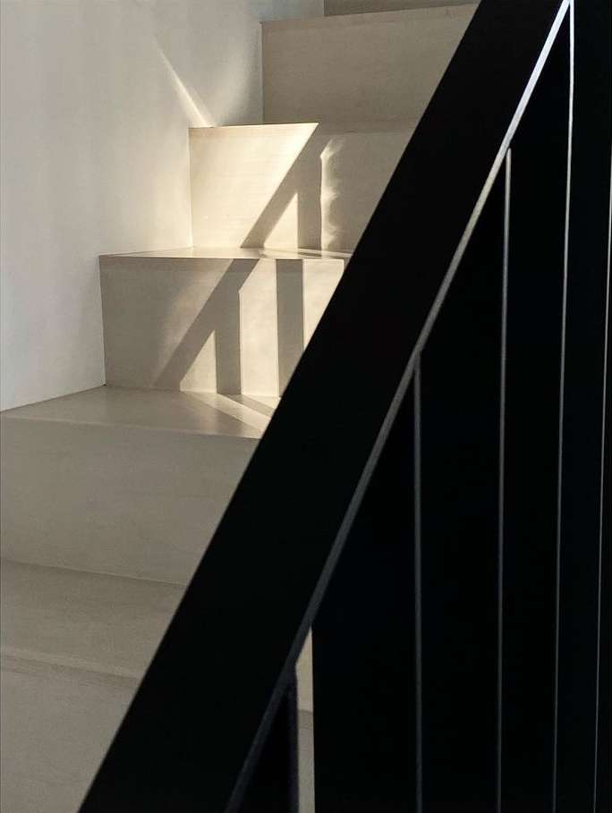 Indoor staircase with a black metal railing, bathed in natural light creating geometric shadows on the stairs and white walls.