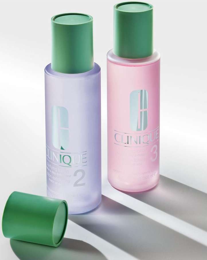 Two Clinique clarifying lotion bottles are standing on a white surface with the open green cap laying beside the bottle on the left.