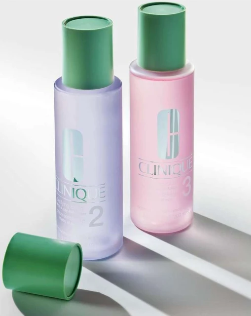 Two Clinique clarifying lotion bottles are standing on a white surface with the open green cap laying beside the bottle on the left.