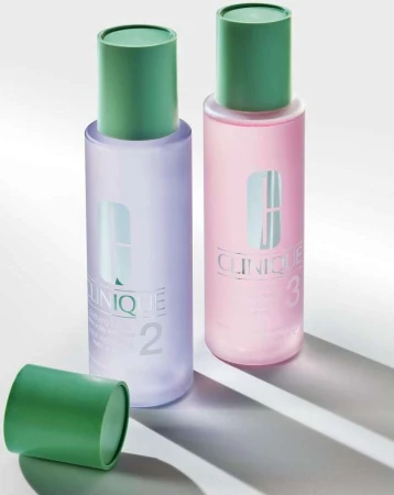 Two Clinique clarifying lotion bottles are standing on a white surface with the open green cap laying beside the bottle on the left.