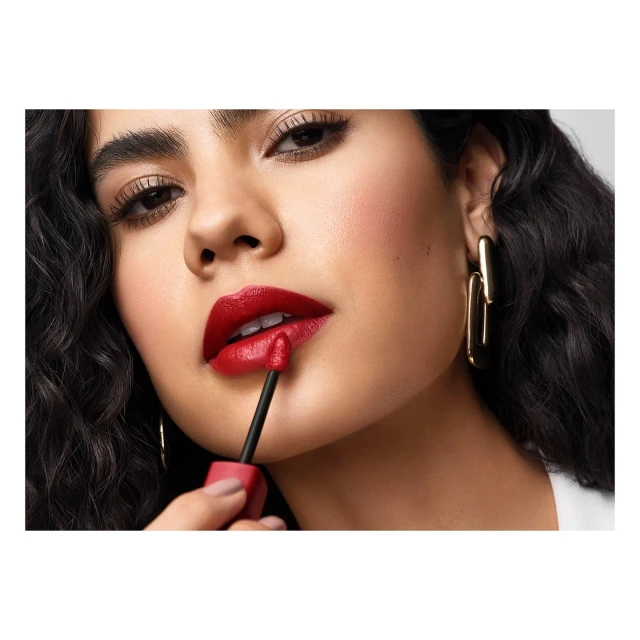 Close-up shot of a woman with dark curly hair applying red lipstick with an applicator brush for beauty or cosmetics advertising.