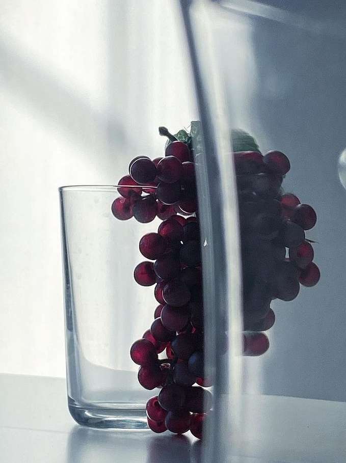 Still life photograph of a glass and cluster of red grapes on a white surface with neutral lighting, emphasizing the simple composition.