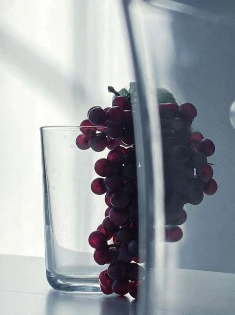 Still life photograph of a glass and cluster of red grapes on a white surface with neutral lighting, emphasizing the simple composition.