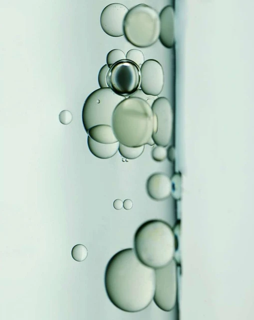 Close-up of translucent liquid with suspended bubbles of varying sizes, some clustered, against a soft, light background.