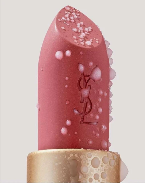 Macro studio shot of a peach-colored lipstick with water droplets and the YSL logo, set against a soft, neutral background.