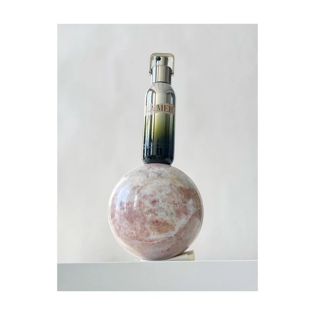 La Mer serum bottle on a rose quartz sphere against a white background. Product shot with luxury skincare focus in an indoor setting.
