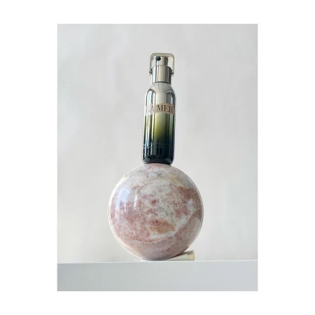 La Mer serum bottle on a rose quartz sphere against a white background. Product shot with luxury skincare focus in an indoor setting.
