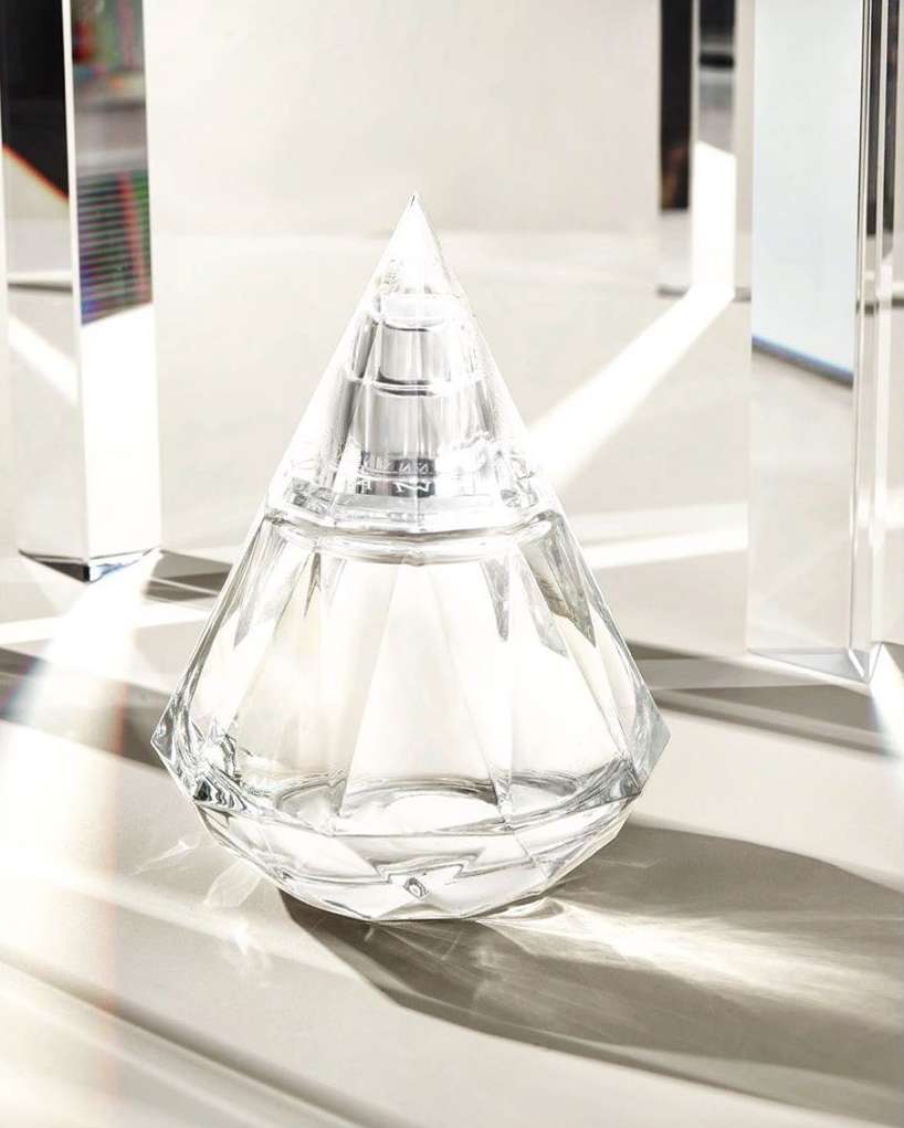 Crystal perfume bottle with diamond-shaped stopper on a shiny white surface with clear geometric objects in the background. Beauty still life.