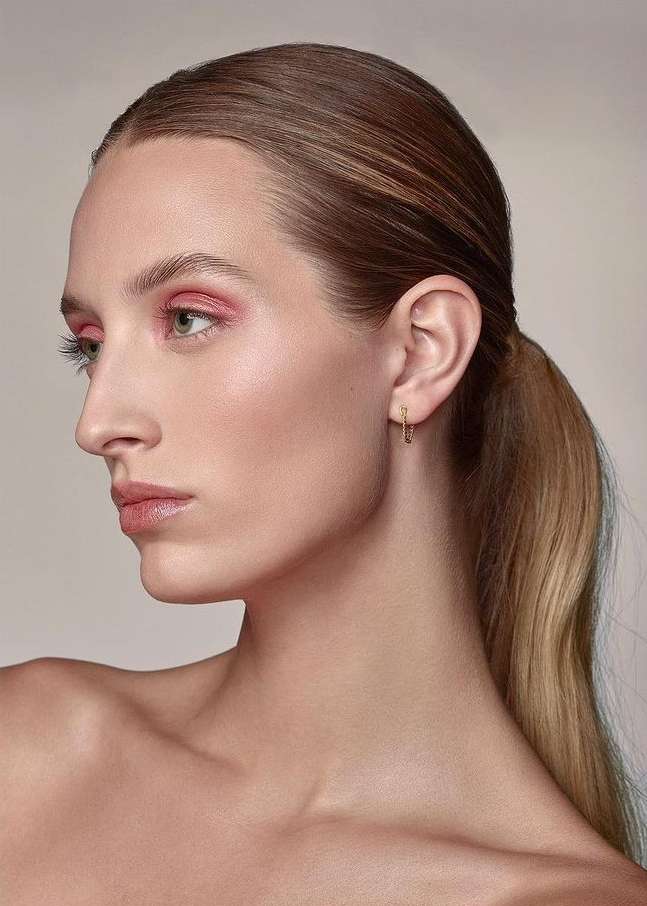 Close-up of an adult woman with pink eyeshadow, gold earrings, and a ponytail against a light background, beauty shot.