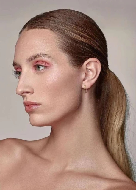 Close-up of an adult woman with pink eyeshadow, gold earrings, and a ponytail against a light background, beauty shot.