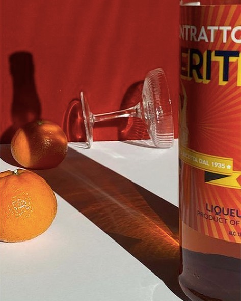Still life of oranges, upturned coupe glass, and a bottle of Contratto Aperitivo against a red background in a studio setting.