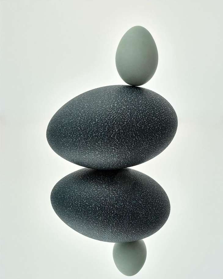 Two stacked eggs, one larger dark blue and one smaller pale blue, balanced on a white reflective surface, studio lighting, minimal composition.