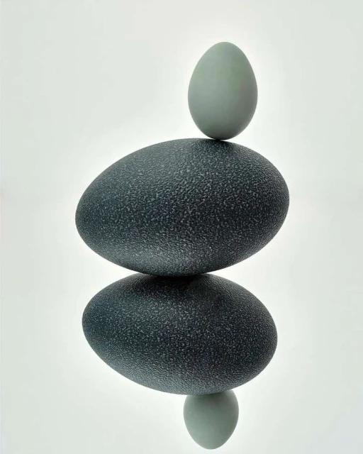 Two stacked eggs, one larger dark blue and one smaller pale blue, balanced on a white reflective surface, studio lighting, minimal composition.