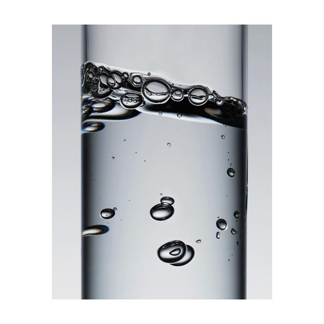 A close-up photo of clear liquid and bubbles inside a transparent glass container on a light gray background, creating a minimalist still life.