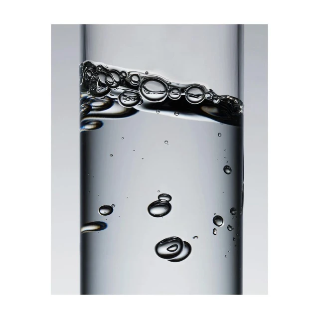 A close-up photo of clear liquid and bubbles inside a transparent glass container on a light gray background, creating a minimalist still life.