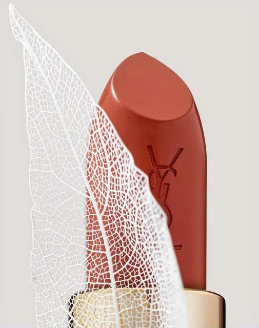 Macro shot of Yves Saint Laurent lipstick in coral color with YSL logo, next to a decorative white leaf with intricate veining.