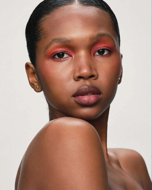 Close-up portrait of a young Black woman with coral makeup on her eyes and cheeks against a neutral background in a beauty shot.