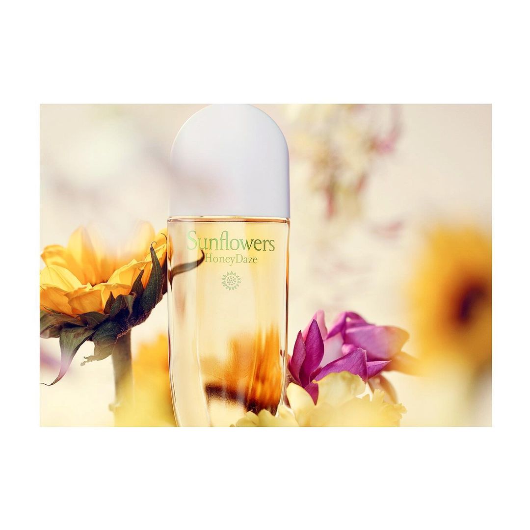 Close-up of a perfume bottle surrounded by yellow and pink flowers, with the brand 'Sunflowers' visible, set against a blurred background.