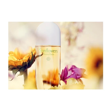 Close-up of a perfume bottle surrounded by yellow and pink flowers, with the brand 'Sunflowers' visible, set against a blurred background.