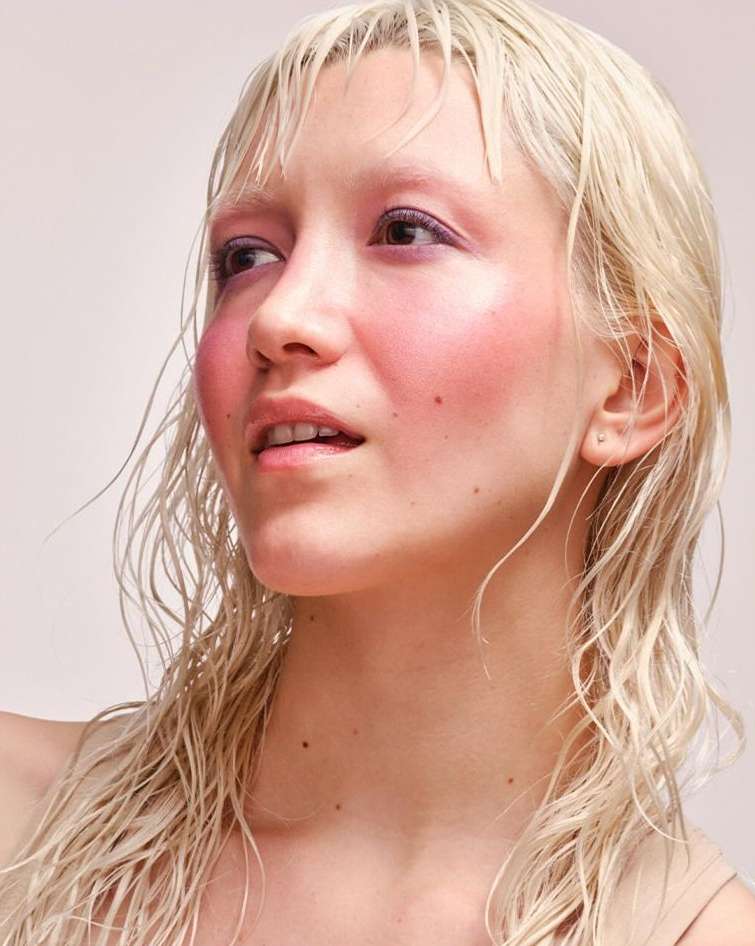 Portrait of a fair-skinned adult female with wet blonde hair, light pink makeup, and a neutral facial expression, against a neutral background.