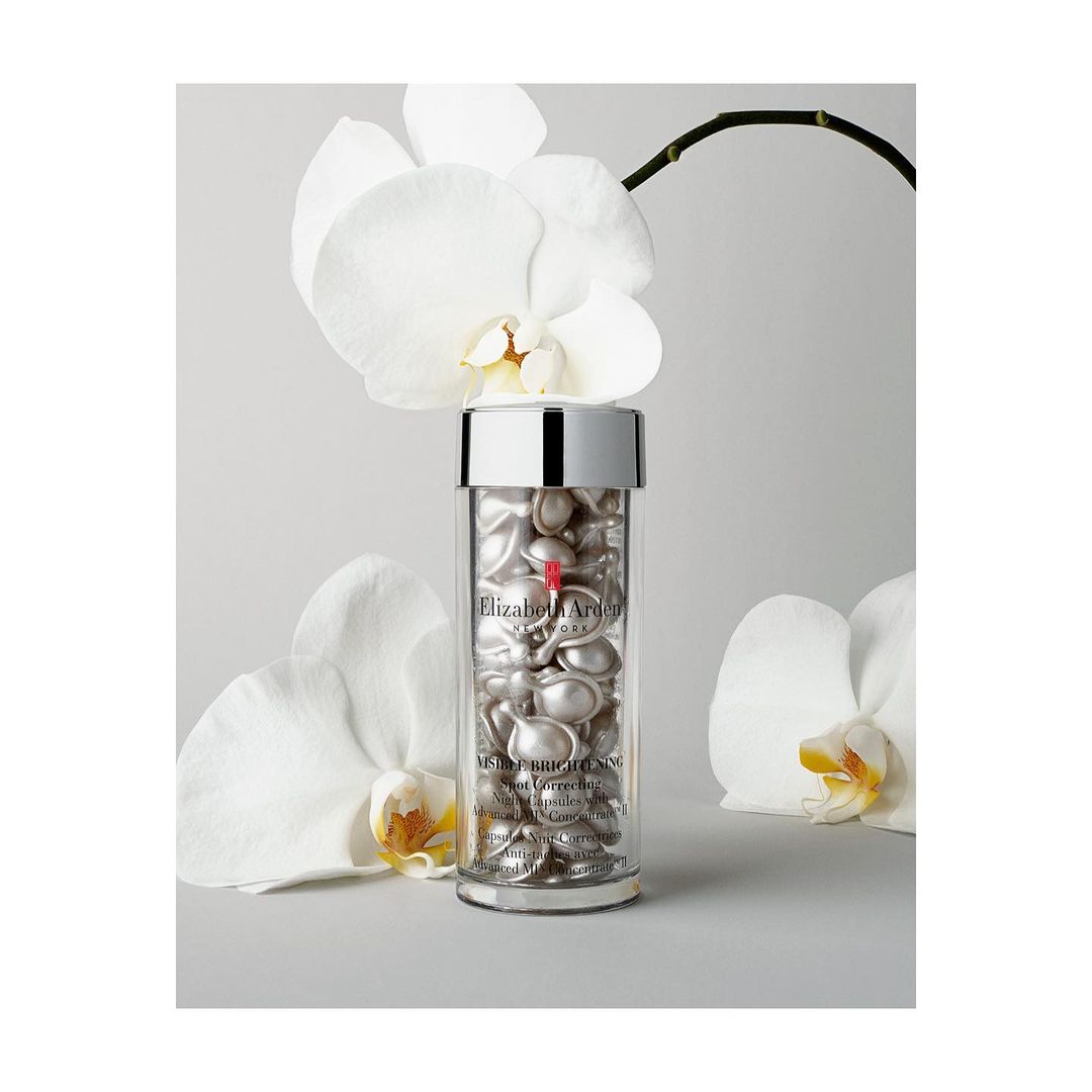 Clear container of Elizabeth Arden Advanced Ceramide capsules surrounded by white orchid blossoms on a light gray surface.