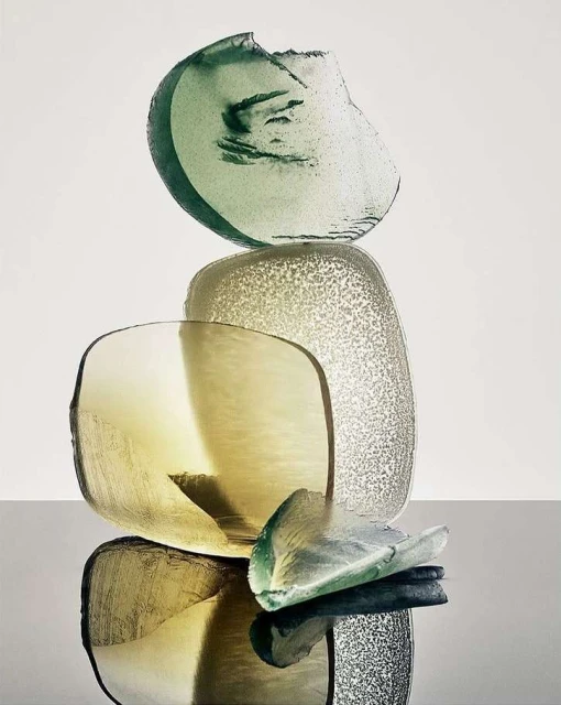 Still life photography featuring stacked green and tan translucent glass shards on a reflective black surface against a white background.