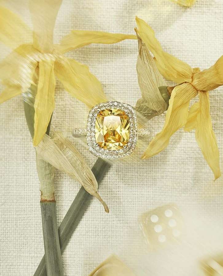Cushion-cut yellow gemstone ring with diamond halo on linen surrounded by yellow flower petals in a jewelry still life.