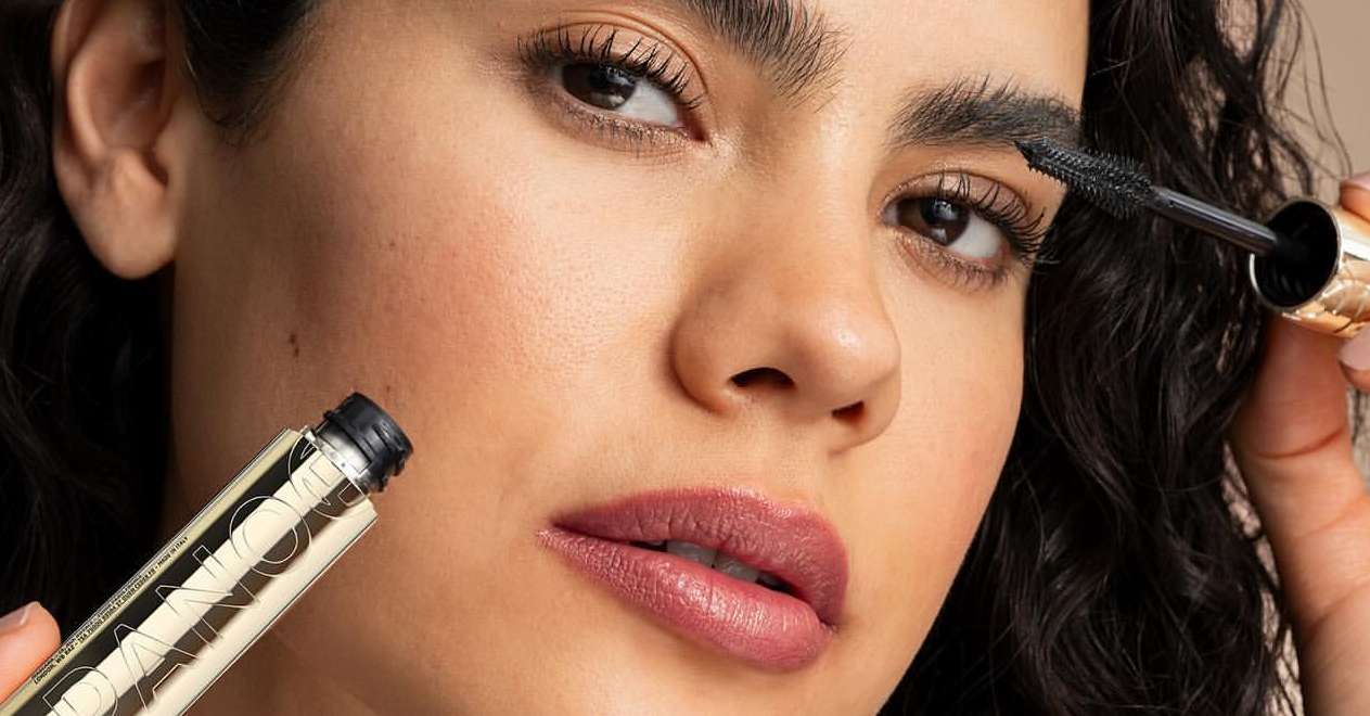 Close-up of a brunette adult female applying mascara to her eyebrow, showcasing beauty and cosmetic product, with a neutral background.