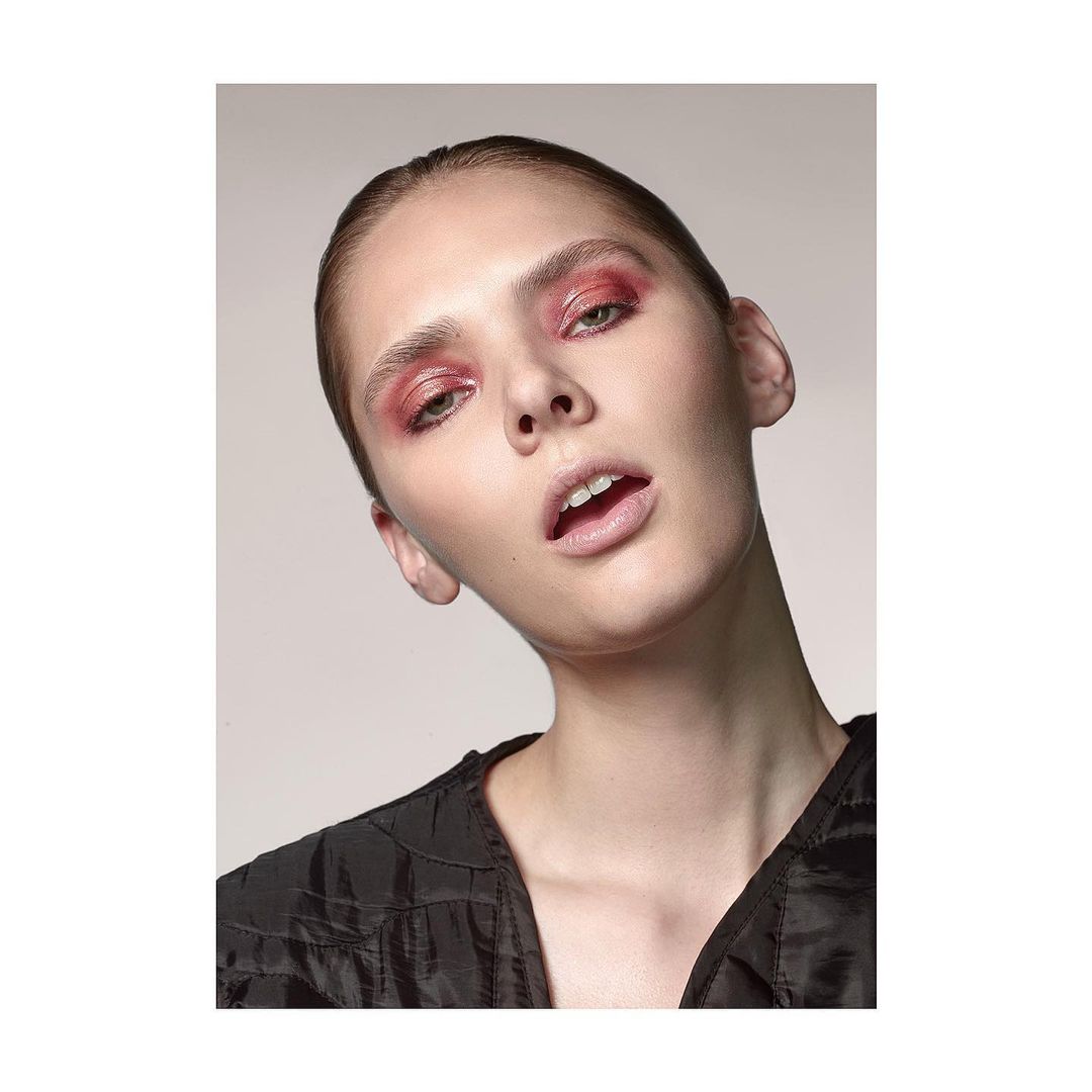 Close-up portrait of a woman with pink eyeshadow and a black textured top against a neutral background.