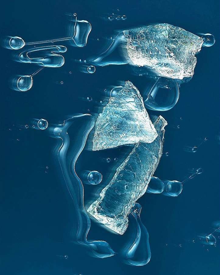 Abstract still life of broken clear ice pieces and water droplets suspended in mid-air against a blue backdrop. Evokes coolness.