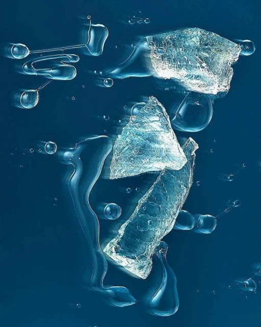 Abstract still life of broken clear ice pieces and water droplets suspended in mid-air against a blue backdrop. Evokes coolness.
