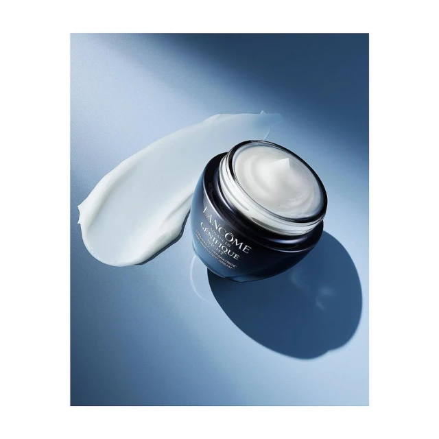 Lancôme Genifique cream in a black jar next to a dab of face cream on a blue background in a studio setting, product shot, beauty ad.