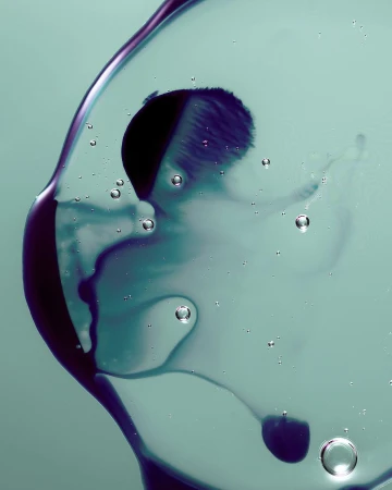 Abstract macro photograph of clear liquid with small bubbles and purple ink splotch against a muted green background, studio shot.