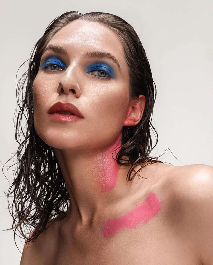 Close-up of a model with wet hair and pink and blue makeup against a white background in a beauty style fashion shoot.