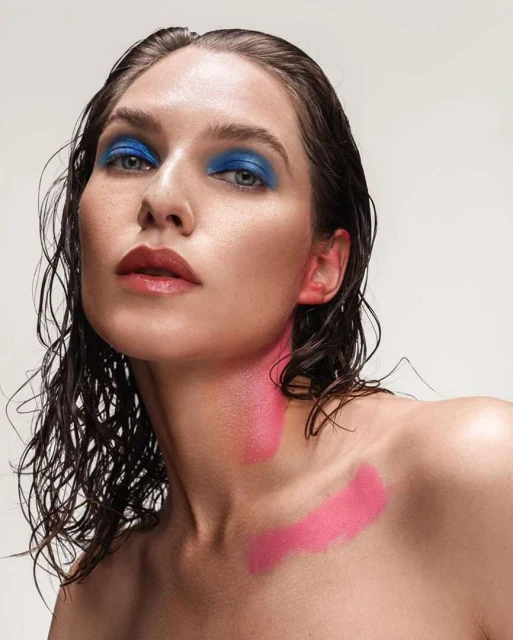 Close-up of a model with wet hair and pink and blue makeup against a white background in a beauty style fashion shoot.