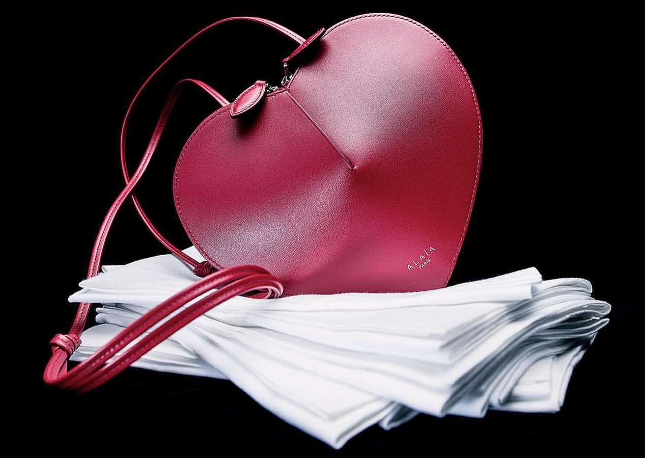 Heart-shaped Alaïa handbag in dark red leather with thin shoulder straps resting on soft, folded white fabric against black background.