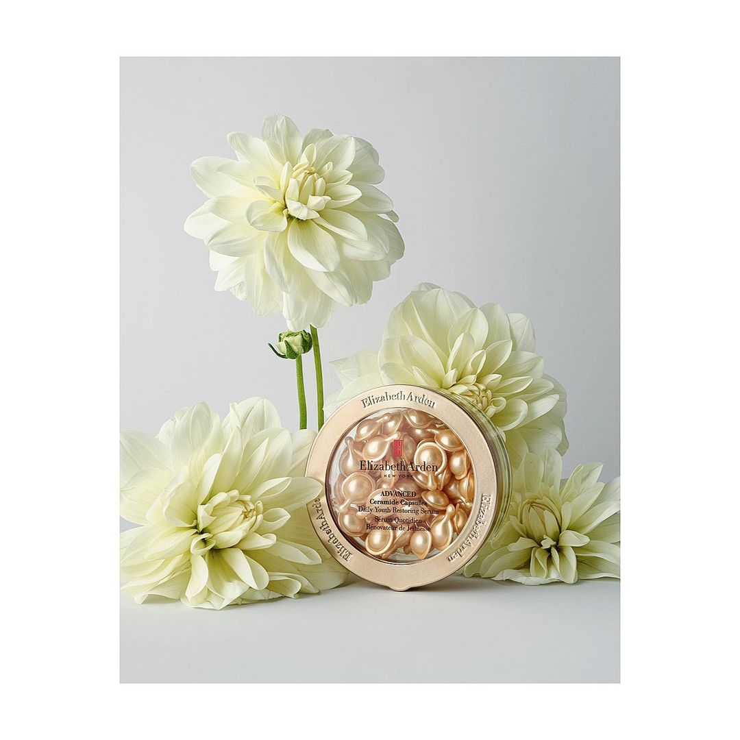 Elizabeth Arden skincare capsules in a gold container, surrounded by several white dahlia flowers against a white backdrop in a studio.
