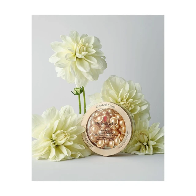 Elizabeth Arden skincare capsules in a gold container, surrounded by several white dahlia flowers against a white backdrop in a studio.