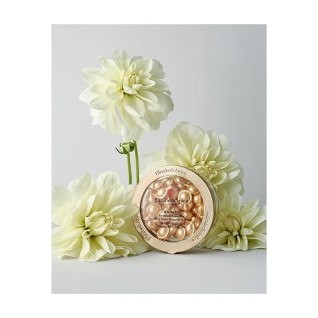 Elizabeth Arden skincare capsules in a gold container, surrounded by several white dahlia flowers against a white backdrop in a studio.