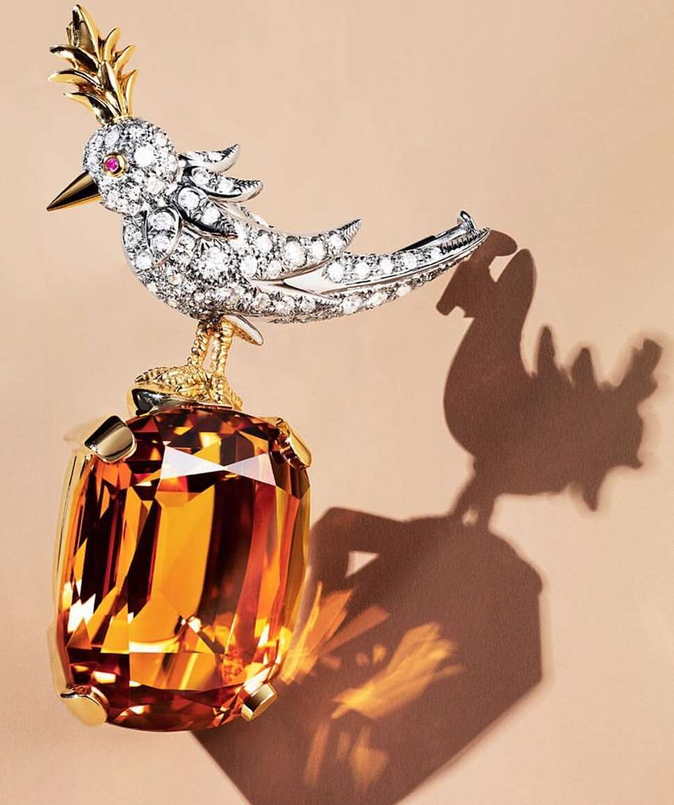 Artistic still life featuring a jeweled bird brooch with diamonds and gold, perched atop a multifaceted orange gemstone on a neutral background.