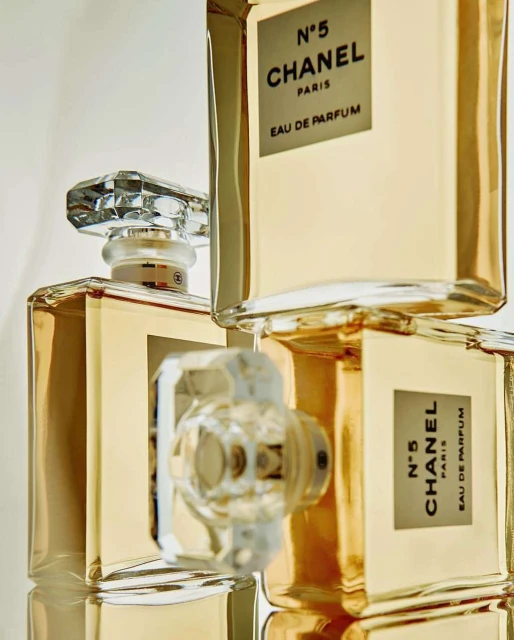 Three Chanel N°5 Eau de Parfum bottles stacked, clear crystal stoppers, gold liquid, white labels with black text, minimalist product photography.